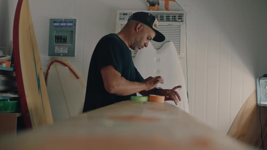 A man in a cap is fixing a surfboard with a tape and then he starts fiddling with his watch