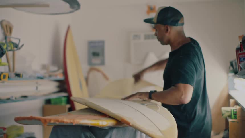 A person peeling off the cover of a a person carefully peels off the cover of a surfboard in a workshop surrounded by various tools and materials