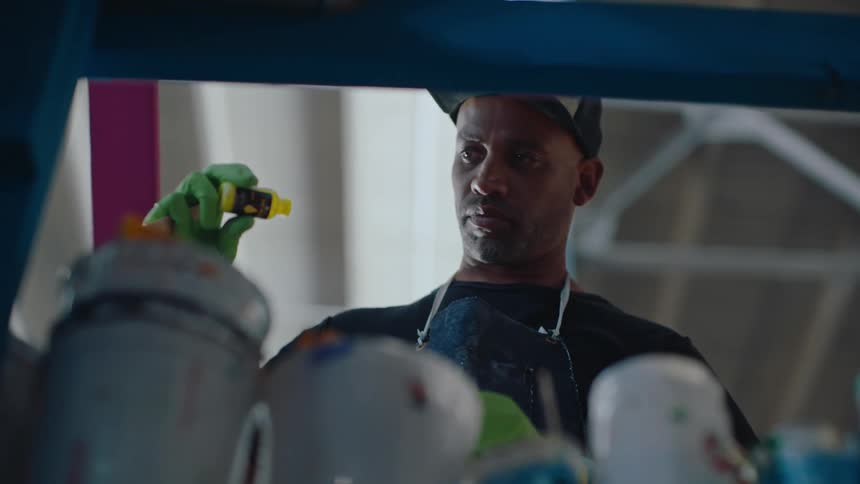 Man wearing a cap and apron handling a small yellow tube of paint