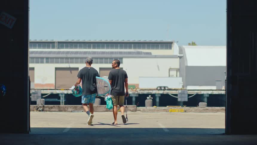 A couple of friends walking out of a warehouse carrying plastic bags and a surfboard