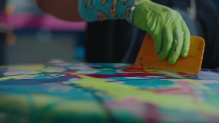Gloved hand holding a spatula passing paint over the colored surface of a surfboard