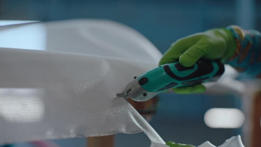 In a workshop a person wearing gloves is cutting a coating material on a surfboard