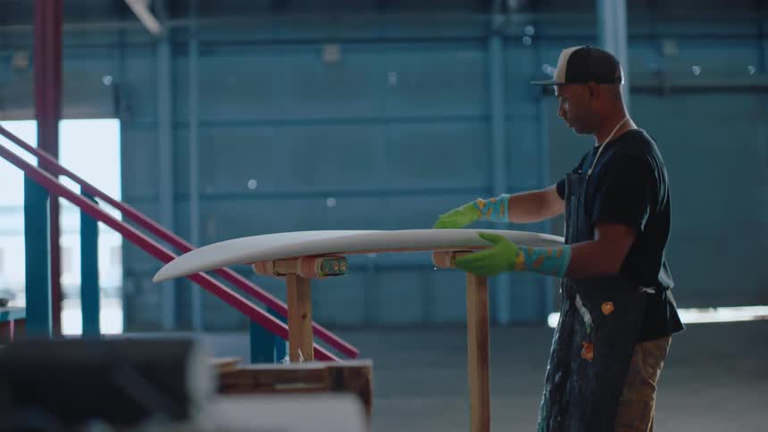 A man with gloves and wearing a cap is placing a surfboard on some wooden pillars