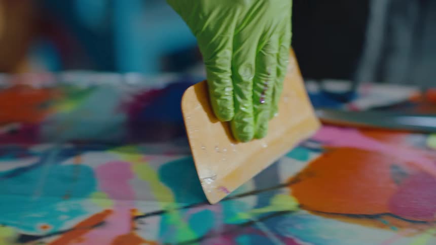 Person removes excess paint from colorful cover on a surfboard in the workshop