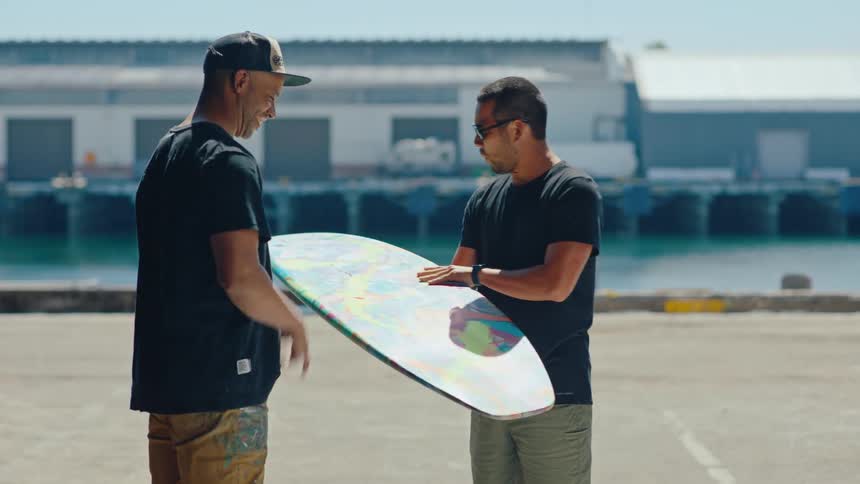A man in a cap handing a colorful surfboard to another man