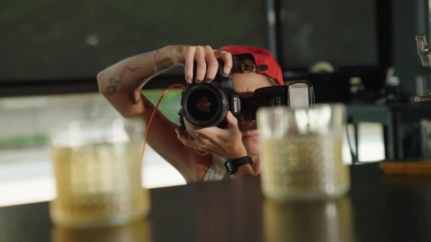A person with a red cap taking a photograph of glasses on a table using a black dslr camera