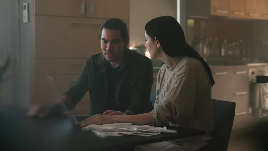 A couple sits at a table near the kitchen talking while focusing on a laptop