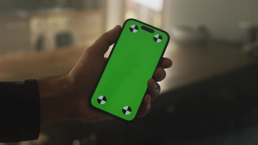A white man holds a smartphone with a green screen while standing in a kitchen