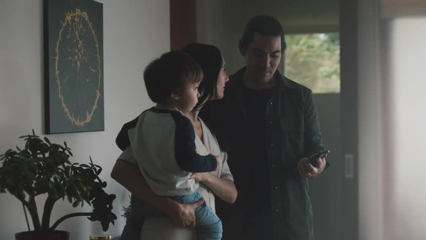 A couple stands on the living room talking to each other while looking at a phone one of them holding a young child