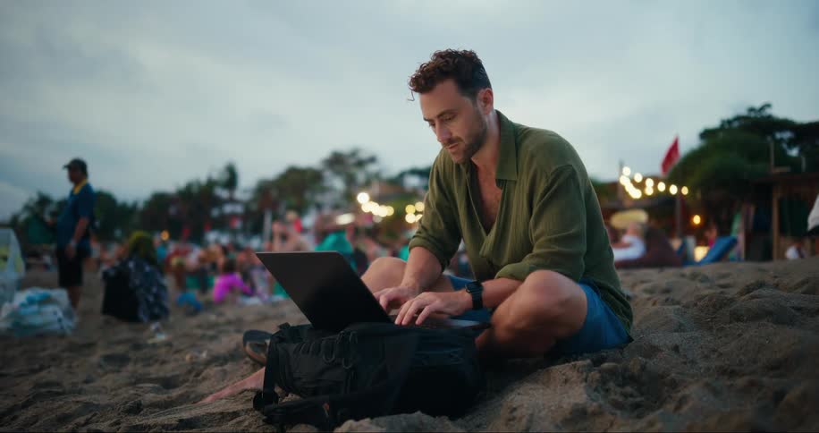 Man sitting on the sand at the beach typing on a laptop and in the background a crowd of people in the late afternoon
