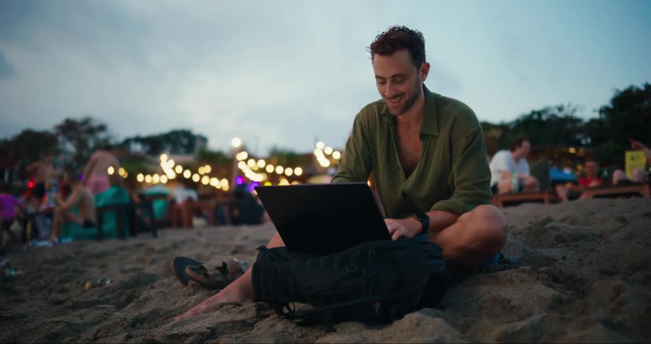 A man sitting on the beach working on a laptop laughing as he types