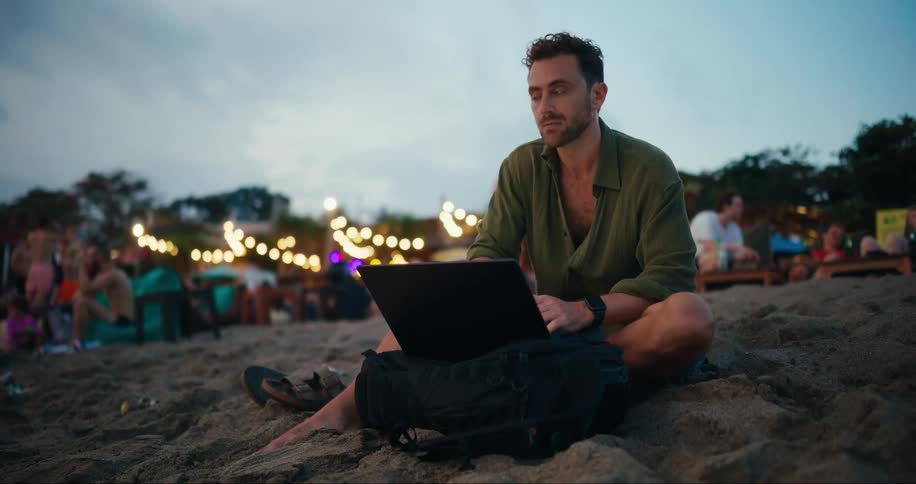 A man sits apart from an outdoor gathering to type on his laptop