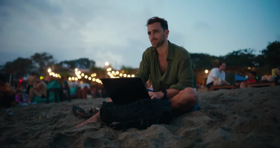 A man sitting on a beach typing on a laptop at dusk