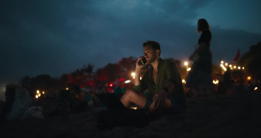 Man sitting on the sand at the beach talking on his cell phone at night with several people around him