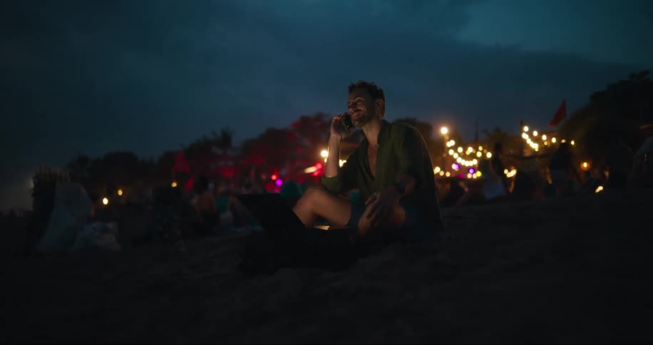 Man sitting on the sand talking on his cell phone with people around and lights in the background in a natural environment at night