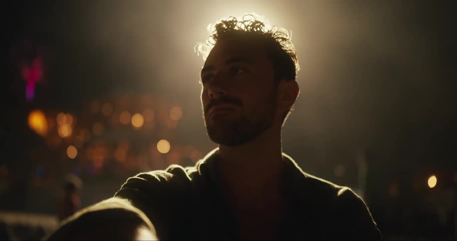 A man sits at an event with mist moving behind him surrounded by several people and lights in the background