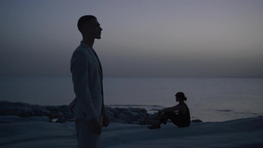 Man in a suit standing and in the background the silhouette of a person sitting by the sea