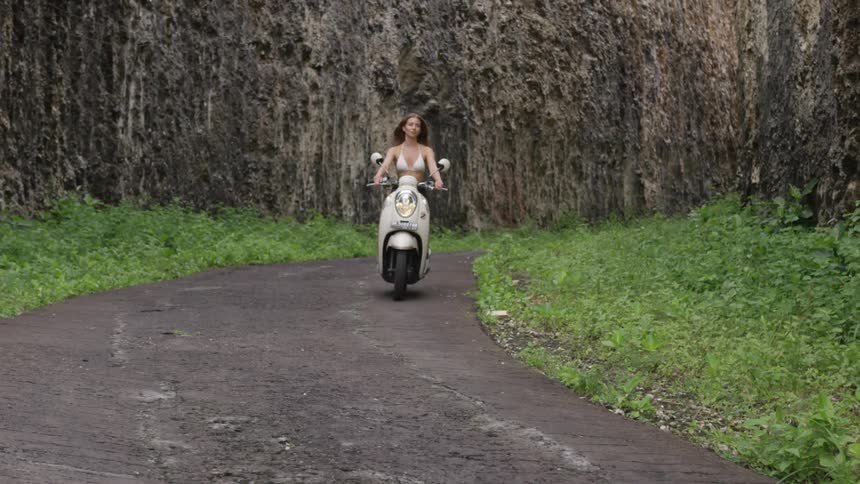 A woman riding a vespa motorcycle on a curved path through a mountain path