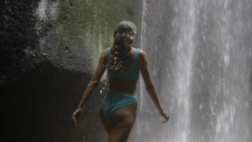 Woman in bikini walking towards a waterfall in a natural setting