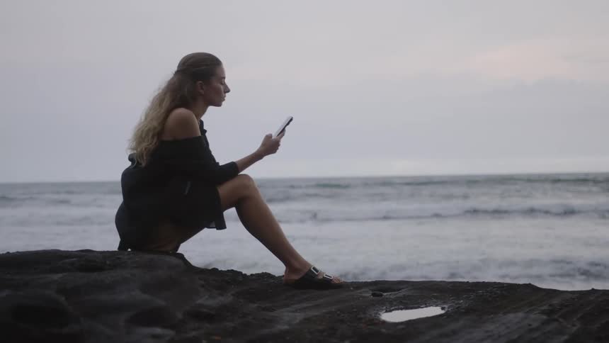 Woman sitting on a rock using a smartphone next to a beach