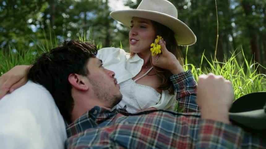 Man lying on a womans lap in a field and placing a yellow flower in her ear