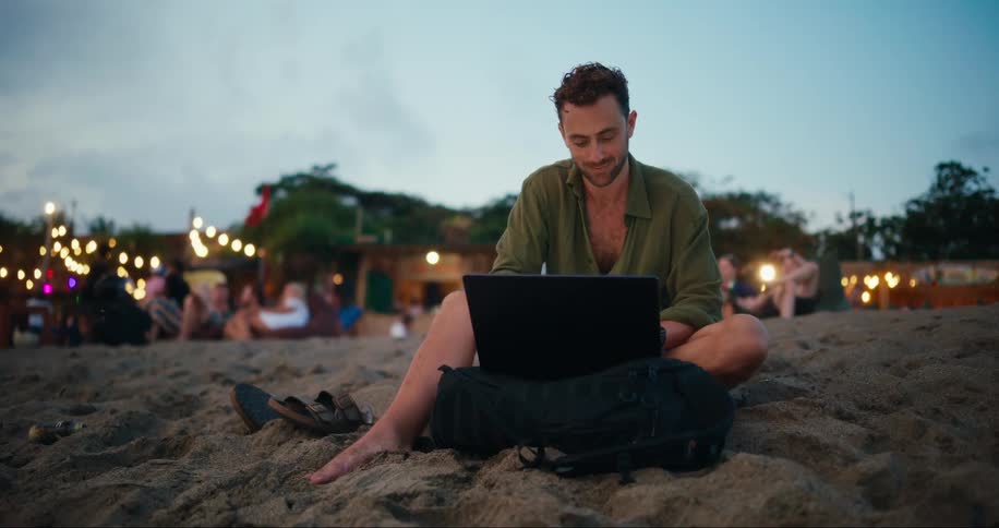 A man is sitting in the sand using his laptop
