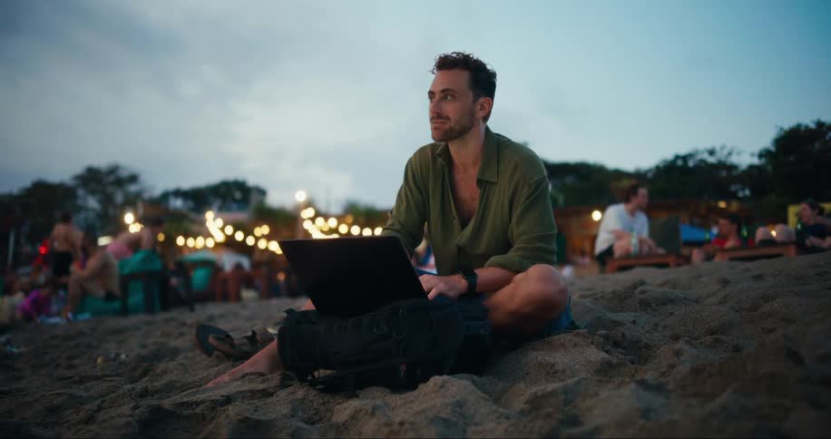 A young man sitting on a sandy beach using a laptop at dusk