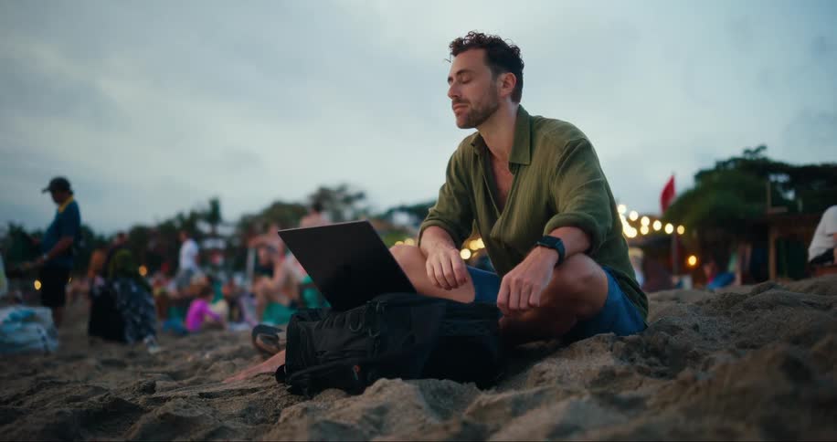 Man sitting on the sand at the beach using a laptop while other people are around him in the late afternoon