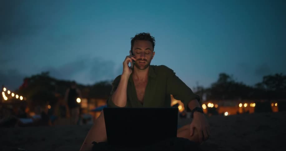 A man is sitting on a beach at dusk engaged in a phone call and smiling while working on a laptop