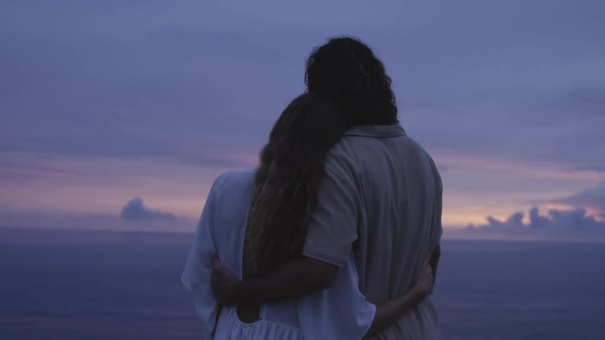 A couple stands by the ocean during sunset seen from behind embracing warmly