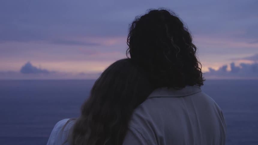 Man and woman standing on the shore cuddle together while watching the sunrise
