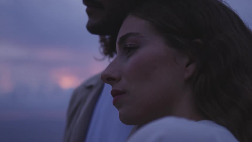 Woman standing next to a man resting her head on his shoulder as they stand in a natural setting against a purple sky