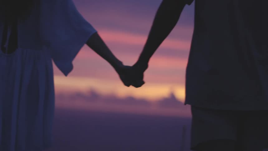 Two people hold hands while walking in a field at sunset