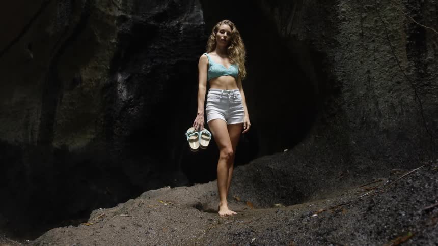 Barefoot woman wearing white shorts holding a sandal in her hands in a natural environment with a rocky wall in the background and a river to the side
