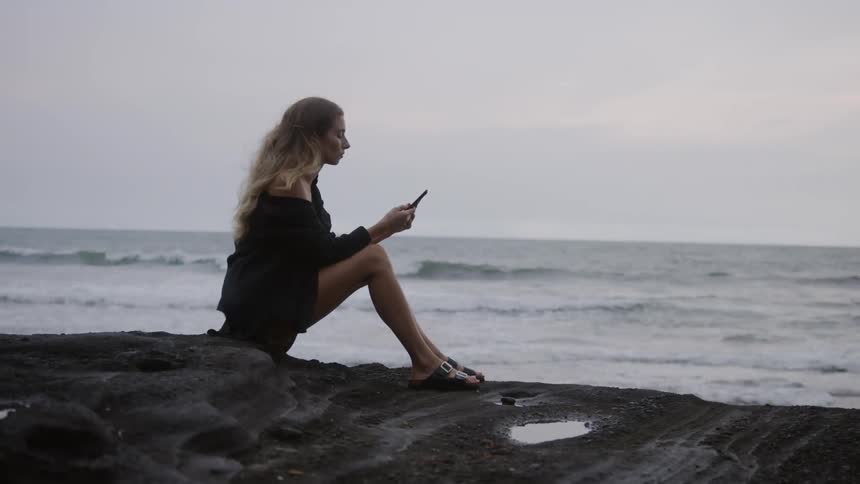 A blonde woman in a black dress is sitting on a rock facing the beach and is using a cell phone