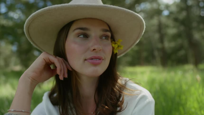 A woman with a flower in her hair wearing a wide brimmed hat sitting thoughtfully in a lush green setting