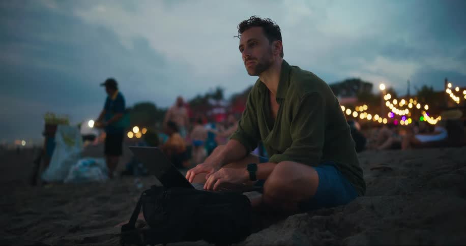 A person sitting on a beach typing on a laptop at dusk