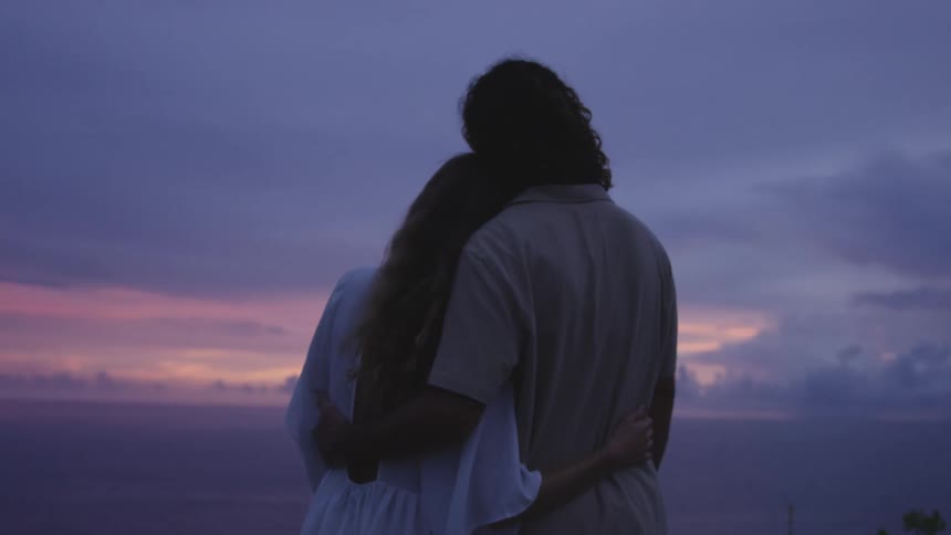 A couple embrace against a pastelcolored sky during sunset