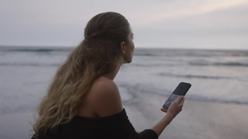 Woman sitting by the sea scrolling through her smartphone screen