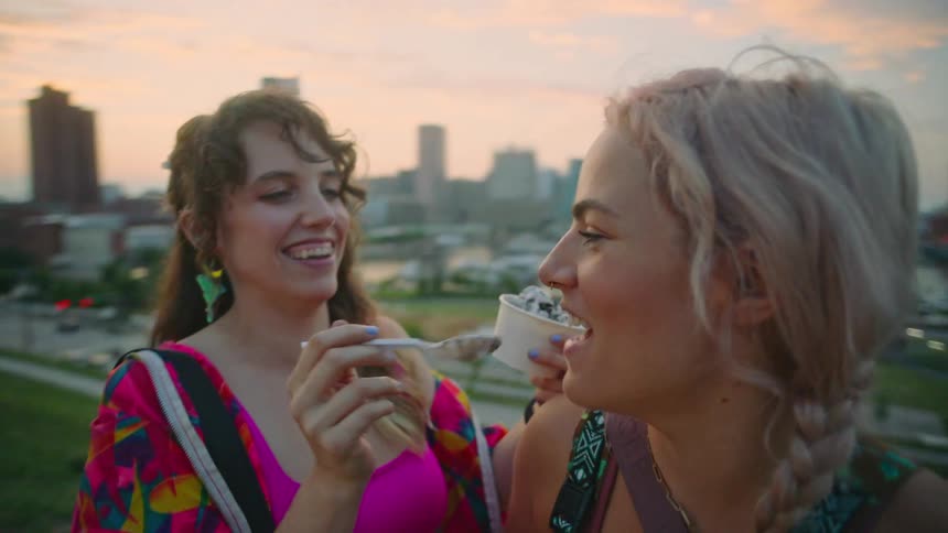 Two female friends laugh while sharing ice cream in a park with a cityscape in the background