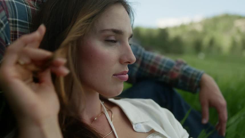 Portrait of a young woman playing with her hair sitting in a field leaning against a man