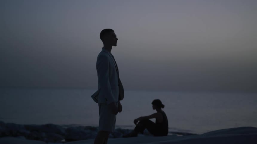 Silhouette of a man standing wearing a blazer and in the background another person sitting by the sea