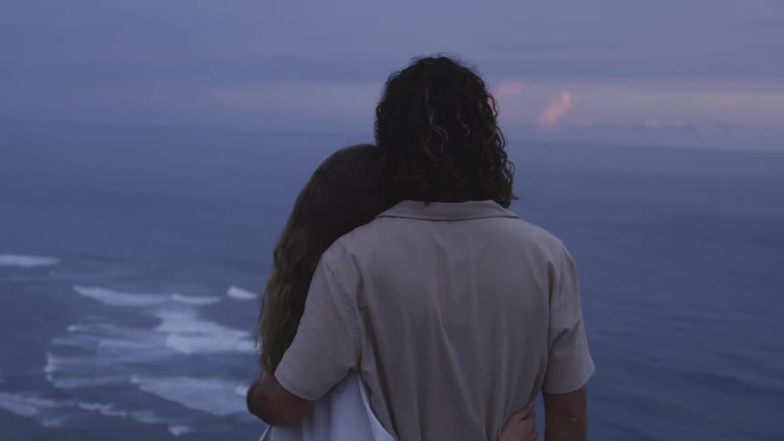 Back view of a woman and a man embracing in an elevated setting by the sea with the sky in shades of purple