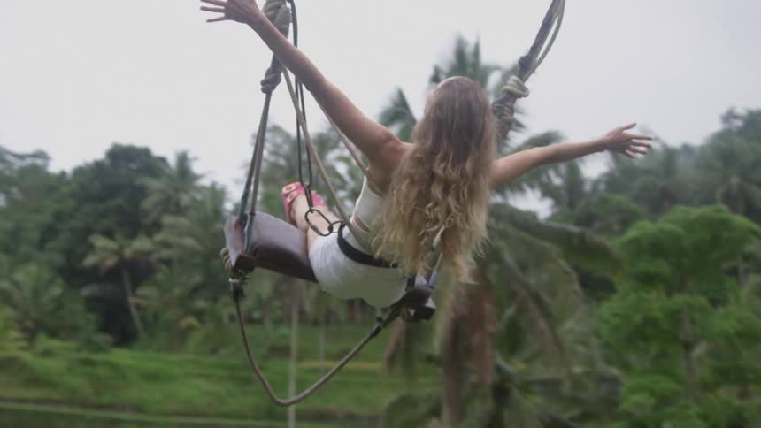 A woman with her arms extended sitting on a giant swing soaring above tropical greenery
