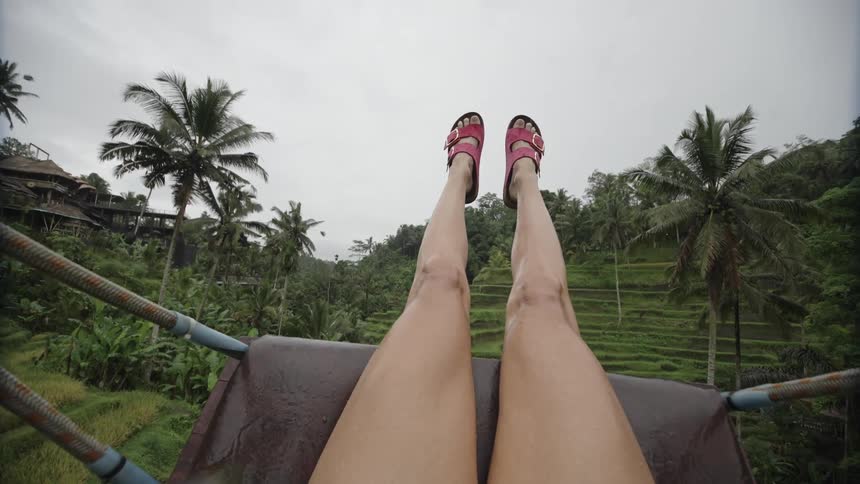 Point of view of a womans legs sitting on a swing in a natural setting with tropical palm trees