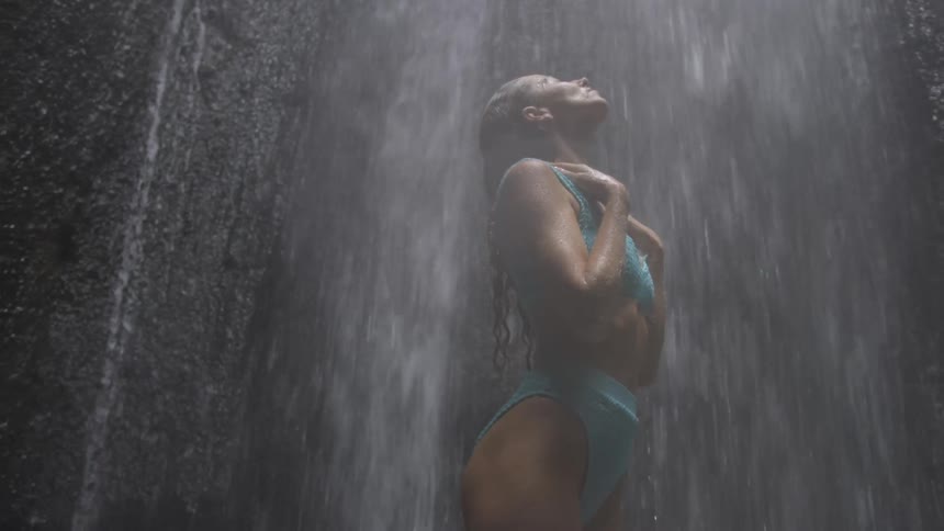 A woman in a blue swimsuit stands under a waterfall
