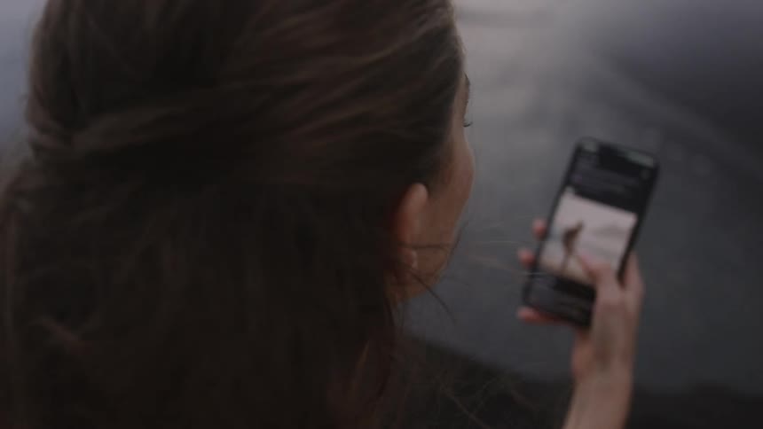 A woman sits on a wet beach scrolling on her smartphone