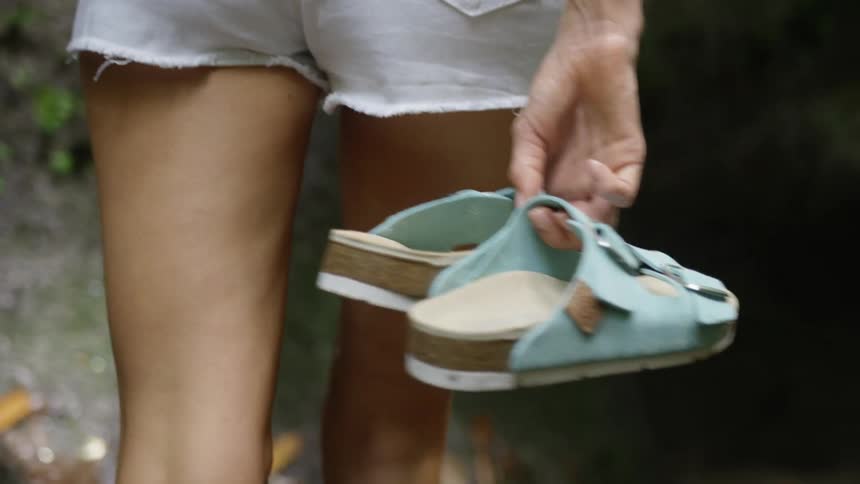 A persons lower legs walk barefoot holding a pair of blue flipflops in one hand