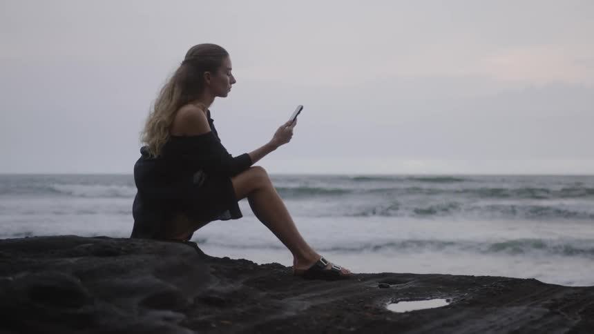 A person sitting on a rock by the sea holding a smartphone during dusk or dawn