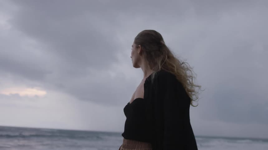 A young blonde woman with long hair walking along the coast looking at the ocean